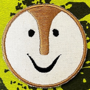 May include: A white embroidered patch with a brown outline, depicting a smiling face with a large brown nose and black eyes.