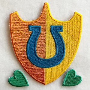 Può includere: Una toppa a forma di scudo gialla e arancione con un ferro di cavallo blu al centro. Due toppe a forma di cuore verde sono su entrambi i lati dello scudo.