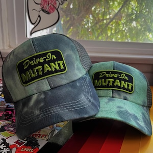 May include: Two green and black trucker hats with a tie-dye pattern. The hats have a black patch with the text "Drive-In Mutant" in green letters.