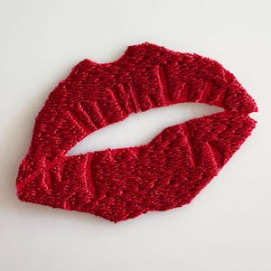 Può includere: Una toppa ricamata rosso brillante a forma di labbra. Il design presenta cuciture dense e strutturate che creano un contorno labiale realistico con un'apertura centrale, mettendo in risalto un vibrante colore di filo cremisi.