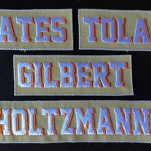 May include: Four beige fabric name tags with white letters outlined in orange. The names are Yates, Tolan, Gilbert, and Holtzman.