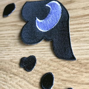 Pu&ograve; includere: Toppa ricamata in nero e viola con un design a forma di luna crescente.