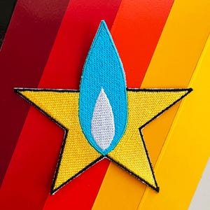 May include: A yellow five-pointed star embroidered patch with a black border. Centered on the star is a vertical, light blue, flame-shaped element containing a smaller, white, teardrop-shaped center, all featuring dense, textured thread stitching.