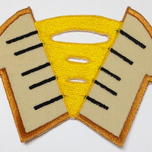 Pode incluir: Um remendo bordado amarelo em forma de sanduíche de queijo grelhado com contorno marrom. O queijo está derretido e tem furos.