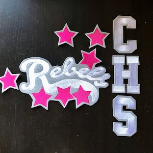 Puede incluir: Parches de fieltro blanco con las palabras "Rebels" y "CHS" en una fuente blanca cursiva. Los parches están decorados con estrellas de fieltro rosa.