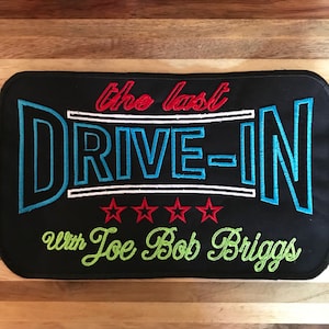 Könnte beinhalten: Schwarzer Stoff-Patch mit weiß und blau gesticktem Text, der "the last DRIVE-IN with Joe Bob Briggs" lautet. Unter dem Text befinden sich fünf rote gestickte Sterne.