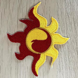 Può includere: Toppa ricamata con un design a forma di sole stilizzato. La toppa è divisa in due metà: una in giallo brillante e l'altra in rosso intenso. Il centro presenta un simbolo yin-yang, anch'esso rosso e giallo.