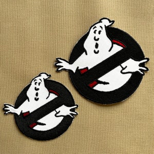 May include: Two embroidered Ghostbusters logo patches. Each patch shows a white ghost figure within a black circle with a diagonal black line. The ghost has red accents, set against a tan background.