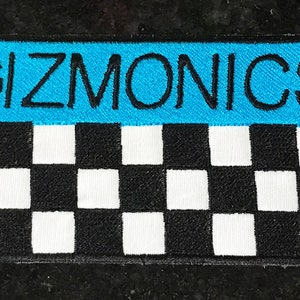 May include: Embroidered patch with the word "GIZMONICS" in black letters on a light blue background. The lower portion features a black and white checkered pattern, creating a visually striking design.