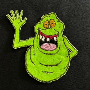May include: Green embroidered patch of a cartoon ghost with a wide open mouth and a toothy grin. The ghost is waving with one arm and has a green body with yellow eyes.