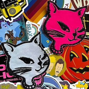 May include: A collage of colorful patches featuring a pink cat with black eyes, a white cat with black eyes, a pumpkin with a jack-o-lantern face, and a variety of other designs. The patches are made of fabric and have a variety of textures.