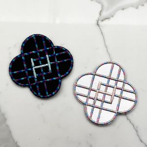Puede incluir: Dos parches bordados en forma de trébol. Uno es negro con un patrón de cuadrícula en verde azulado, azul y rosa, y una letra "H" blanca. El otro es blanco con un patrón de cuadrícula similar y una "H" rosa.