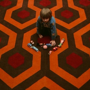 May include: A young child in a blue sweater sits on a red and orange patterned carpet. The child is playing with toy cars and a pink ball.
