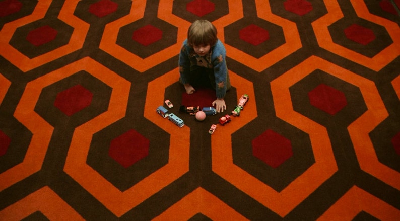 May include: A young child in a blue sweater sits on a red and orange patterned carpet. The child is playing with toy cars and a pink ball.