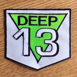 Puede incluir: Un parche blanco con un borde negro en forma de escudo. Dentro del escudo hay un triángulo verde con el texto "DEEP" en negro y el número "13" en blanco.