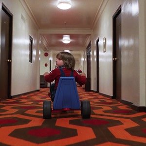 May include: A young boy in a red shirt and blue overalls rides a blue tricycle down a long hallway with a patterned carpet. The carpet is orange and brown with a repeating geometric pattern.