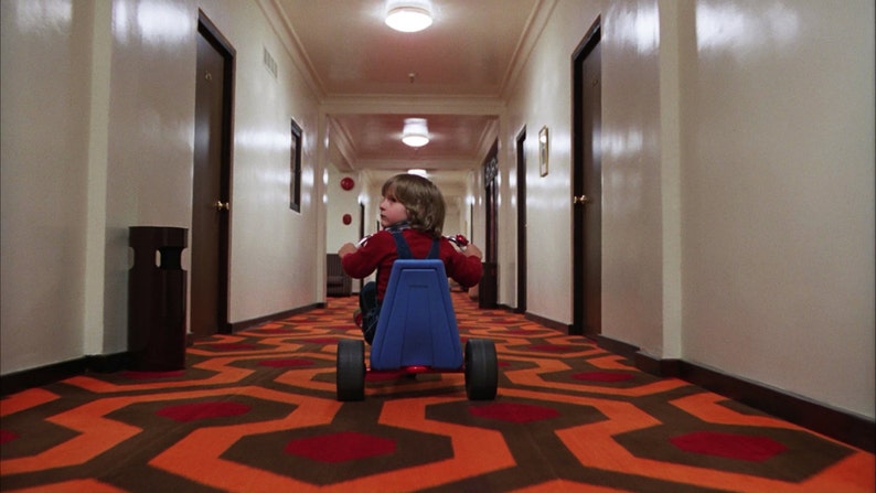 May include: A young boy in a red shirt and blue overalls rides a blue tricycle down a long hallway with a patterned carpet. The carpet is orange and brown with a repeating geometric pattern.