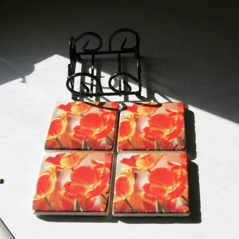 Op de afbeelding: Vier vierkante keramische onderzetters met een bloemenontwerp met oranje tulpen op een witte achtergrond. De onderzetters worden gepresenteerd in een zwart metalen rek.