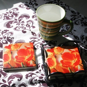 Op de afbeelding: Vier onderzetters met oranje en gele tulpenmotieven in een zwart metalen frame. De onderzetters liggen op een wit en bruin gepatroneerd tafelkleed.