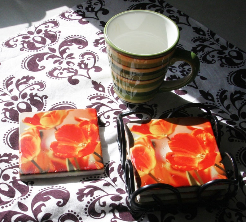 Op de afbeelding: Vier onderzetters met oranje en gele tulpenmotieven in een zwart metalen frame. De onderzetters liggen op een wit en bruin gepatroneerd tafelkleed.