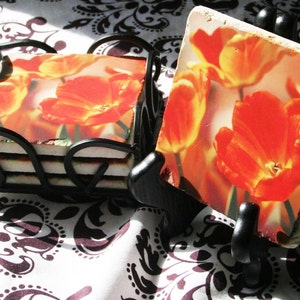 Op de afbeelding: Set van vier vierkante keramische onderzetters met een bloemenontwerp met oranje tulpen. De onderzetters worden gepresenteerd op een zwarte metalen standaard.