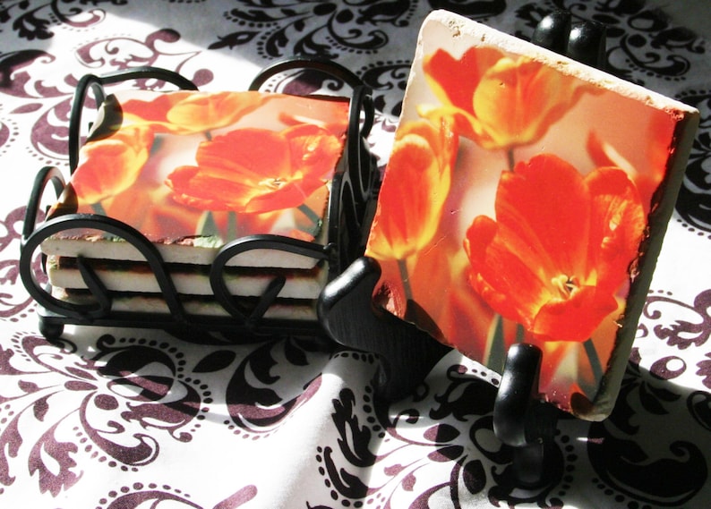 Op de afbeelding: Set van vier vierkante keramische onderzetters met een bloemenontwerp met oranje tulpen. De onderzetters worden gepresenteerd op een zwarte metalen standaard.