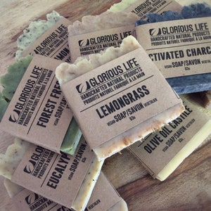 Puede incluir: Surtido de jabones naturales hechos a mano con varios aromas, incluyendo Lemongrass, Bosque Fresco, Eucalipto, Carb&oacute;n Activado y Aceite de Oliva de Castilla. Cada barra est&aacute; etiquetada con el aroma y las palabras "Glorious Life" y "Jab&oacute;n Vegano".