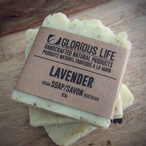 Puede incluir: Tres barras de jab&oacute;n de lavanda hecho a mano envueltas en papel marr&oacute;n con el texto "GLORIOUS LIFE HANDCRAFTED NATURAL PRODUCTS PRODUITS NATUREL FABRIQU&Eacute; &Agrave; LA MAIN LAVENDER VEGAN SOAP/SAVON VEGETALIEN 63g" impreso en la etiqueta.