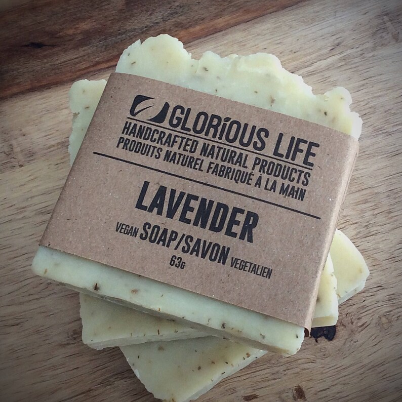 Puede incluir: Tres barras de jab&oacute;n de lavanda hecho a mano envueltas en papel marr&oacute;n con el texto "GLORIOUS LIFE HANDCRAFTED NATURAL PRODUCTS PRODUITS NATUREL FABRIQU&Eacute; &Agrave; LA MAIN LAVENDER VEGAN SOAP/SAVON VEGETALIEN 63g" impreso en la etiqueta.