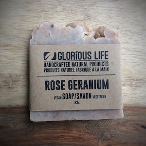 May include: A bar of brown and pink soap with a brown paper label that reads "GLORIOUS LIFE HANDCRAFTED NATURAL PRODUCTS PRODUITS NATUREL FABRIQUÉ À LA MAIN ROSE GERANIUM VEGAN SOAP/SAVON VEGETALIEN 63g".