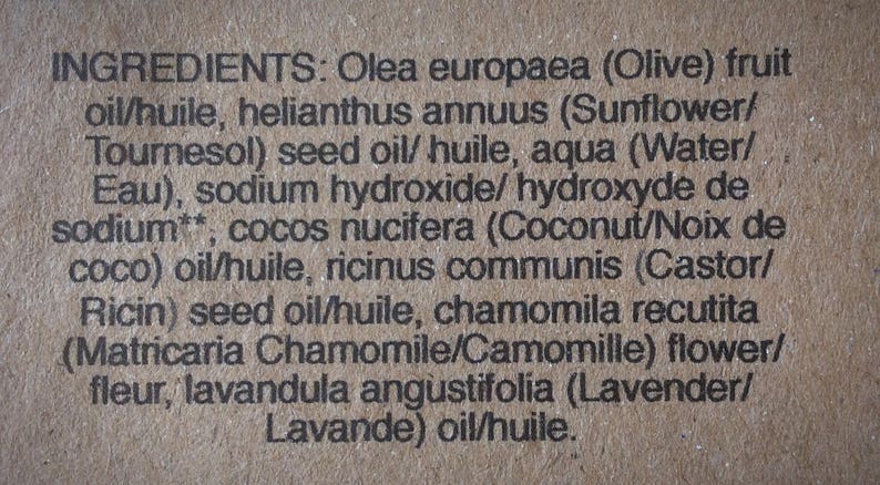 Puede incluir: Un primer plano de una etiqueta de producto que enumera los ingredientes de una pastilla de jab&oacute;n. Los ingredientes se enumeran en ingl&eacute;s y franc&eacute;s. Los ingredientes incluyen aceite de oliva, aceite de semilla de girasol, agua, hidr&oacute;xido de sodio, aceite de coco, aceite de ricino, flor de manzanilla y aceite de lavanda.