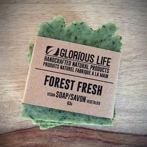 May include: A green, leaf-shaped vegan soap bar with a brown paper label. The label reads "GLORIOUS LIFE FOREST FRESH" and "HANDCRAFTED NATURAL PRODUCTS". The soap weighs 63g. The soap is on a wooden surface.