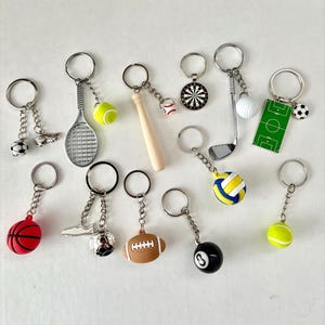 May include: A selection of sports-themed keyrings. Keyrings include a tennis racket with a yellow ball, a baseball bat with a baseball, a golf club with a golf ball, and a football. Other keyrings feature a soccer ball, basketball, and an 8-ball.