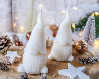 May include: Two white gnome-shaped candles with pointy hats on a wooden surface with pine cones and white stars. The candles are surrounded by a festive winter scene with white lights and greenery.