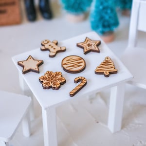 Könnte beinhalten: Ein weißer Tisch mit acht hölzernen Weihnachtsornamenten. Die Ornamente sind in Form eines Lebkuchenmanns, eines Sterns, einer Schneeflocke, eines Zuckerstangs, eines Weihnachtsbaums und eines Kreises.