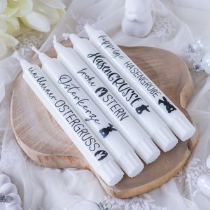 Könnte beinhalten: Sechs weiße Kerzen mit schwarzem Text und Hasenillustrationen. Die Kerzen sind beschriftet mit "Frohe Ostern", "Osterkerze", "Ostergruss", "Hasengruesse", "Hasengrube" und "Hüpflige Hasengrube".