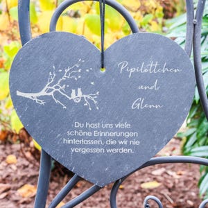 Può includere: Una targa in ardesia nera a forma di cuore con il testo "Pipilottchen und Glenn" e un piccolo albero con due uccelli su un ramo. Il testo sotto i nomi dice "Du hast uns viele schöne Erinnerungen hinterlassen, die wir nie vergessen werden."