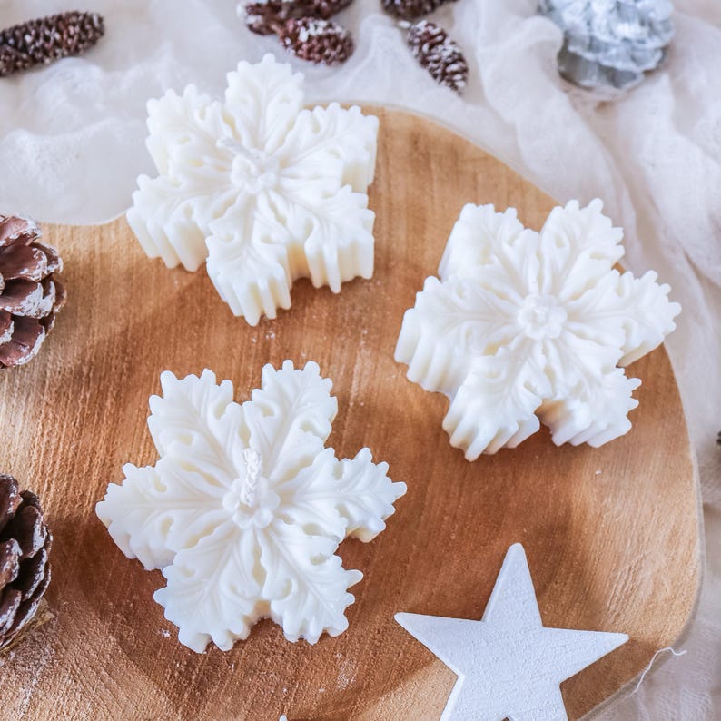 May include: Three white snowflake-shaped candles on a wooden surface. The candles are detailed with intricate designs and are ready to be lit.