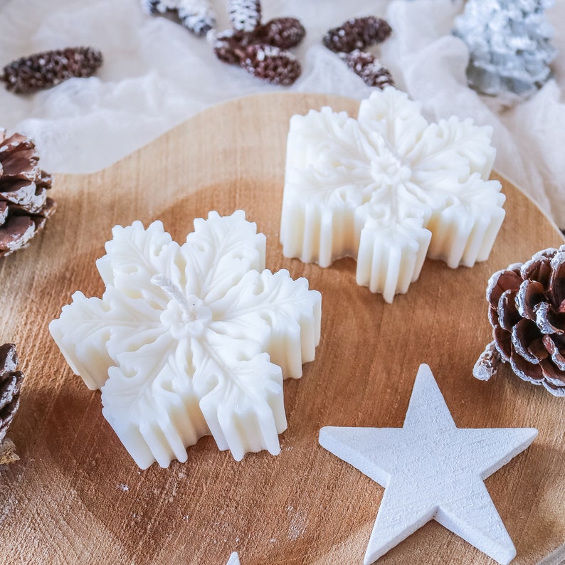 May include: Two white snowflake-shaped candles on a wooden surface. The candles are detailed with intricate designs and have a white star-shaped ornament beside them.
