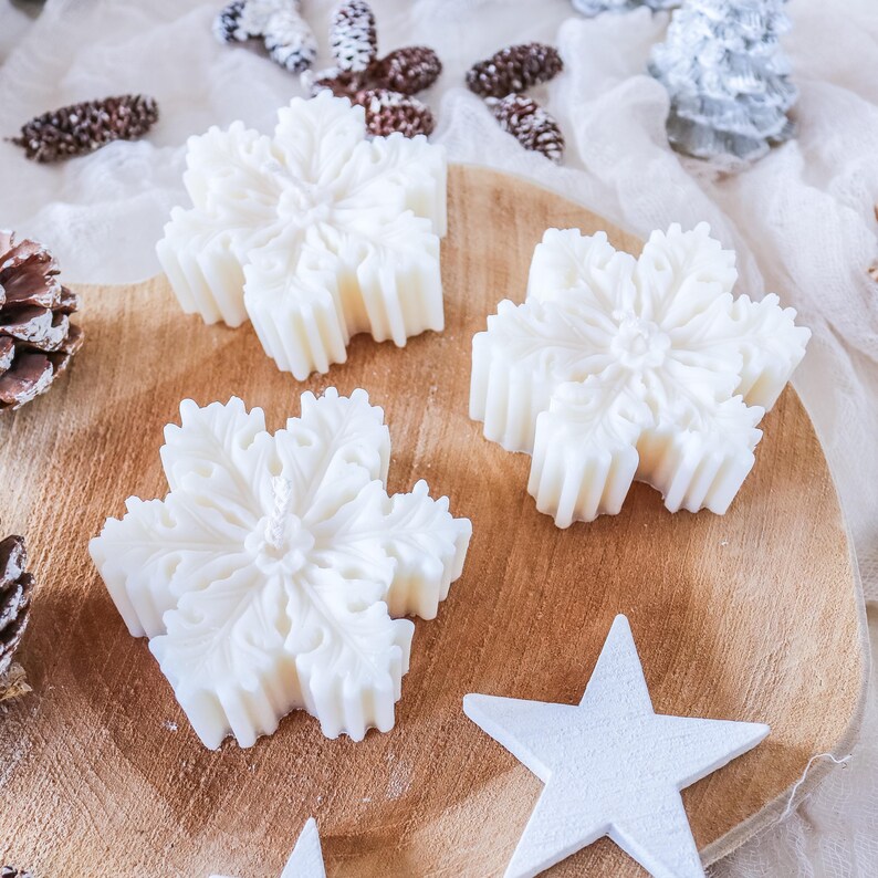 May include: Three white snowflake-shaped candles on a wooden surface. The candles are detailed with intricate designs and are arranged in a triangular pattern.