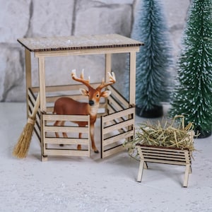 Könnte beinhalten: Ein kleiner Stall aus Holz mit offener Vorderseite, in dem sich eine braune Hirschfigur mit Geweih befindet. Rechts steht eine kleine hölzerne Futterraufe mit trockenem Gras, und ein Miniaturbesen lehnt an der Seite des Stalls.