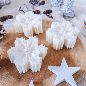 May include: Four white snowflake-shaped candles on a wooden surface. The candles are arranged in a semi-circle with a white star in the center.