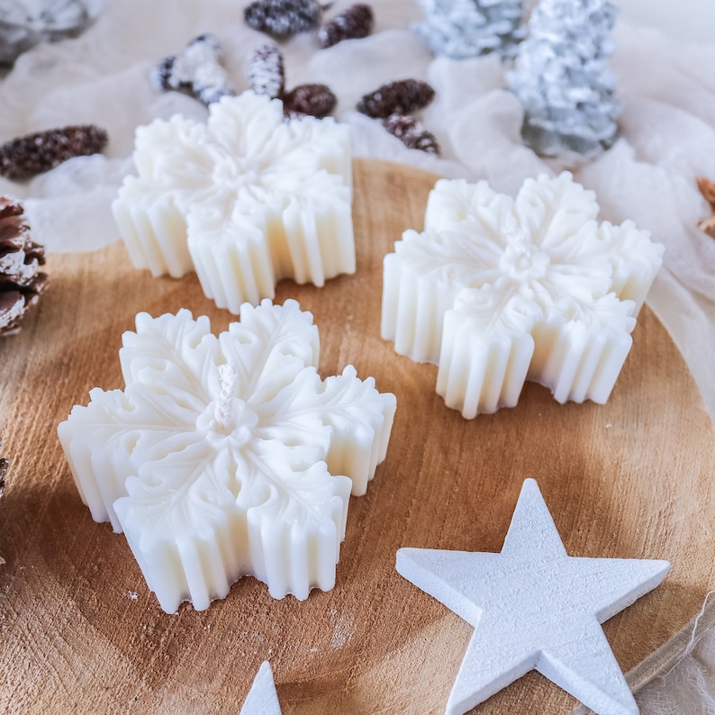 May include: Four white snowflake-shaped candles on a wooden surface. The candles are arranged in a semi-circle with a white star in the center.