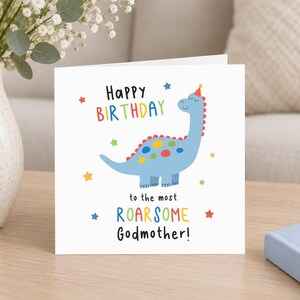 Op de afbeelding: Een vierkante witte wenskaart met een blauwe dinosaurus die een feesthoedje draagt. De tekst luidt Happy Birthday to the most ROARSOME Godmother! Het ontwerp bevat kleurrijke stippen op de dinosaurus en kleine sterretjes.