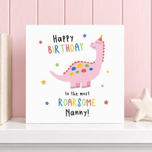 May include: A square greeting card with a white background featuring a pink dinosaur wearing a party hat. The text reads Happy Birthday to the most ROARSOME Nanny! surrounded by colorful stars in red, blue, green, and yellow hues.