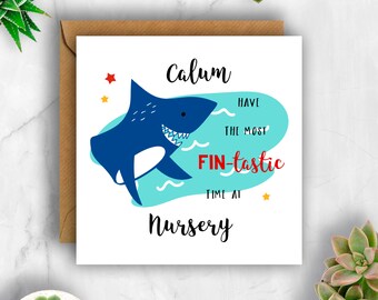 May include: A square greeting card with a cartoon shark illustration. The shark is blue with white teeth and is swimming in a turquoise sea. The card reads "Calum have the most FIN-tastic time at Nursery."