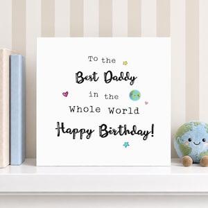 May include: A square white greeting card with black text that reads To the Best Daddy in the Whole World Happy Birthday. The card features small colorful illustrations of a smiling planet Earth, a pink heart, and yellow and blue stars.