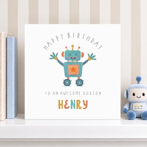 Op de afbeelding: Een vierkante witte wenskaart met een illustratie van een blauwgroene robot met een oranje ster op de borst. De tekst luidt HAPPY BIRTHDAY TO AN AWESOME GODSON HENRY in zwarte en kleurrijke letters. De kaart staat op een witte plank.