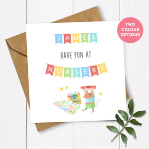 May include: A white card with the text "JAMES HAVE FUN AT NURSERY" in colourful letters. The card features a cartoon mouse holding a crayon and a drawing of a crayon box and a star.