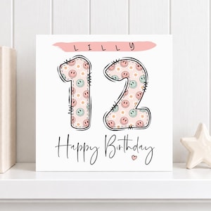 Op de afbeelding: Een vierkante verjaardagskaart met de naam LILLY in zwarte letters op een roze penseelstreek. Het getal 12 is weergegeven in een groot lettertype in stikselstijl, gevuld met een patroon van roze en groene smileys en witte madeliefjes.